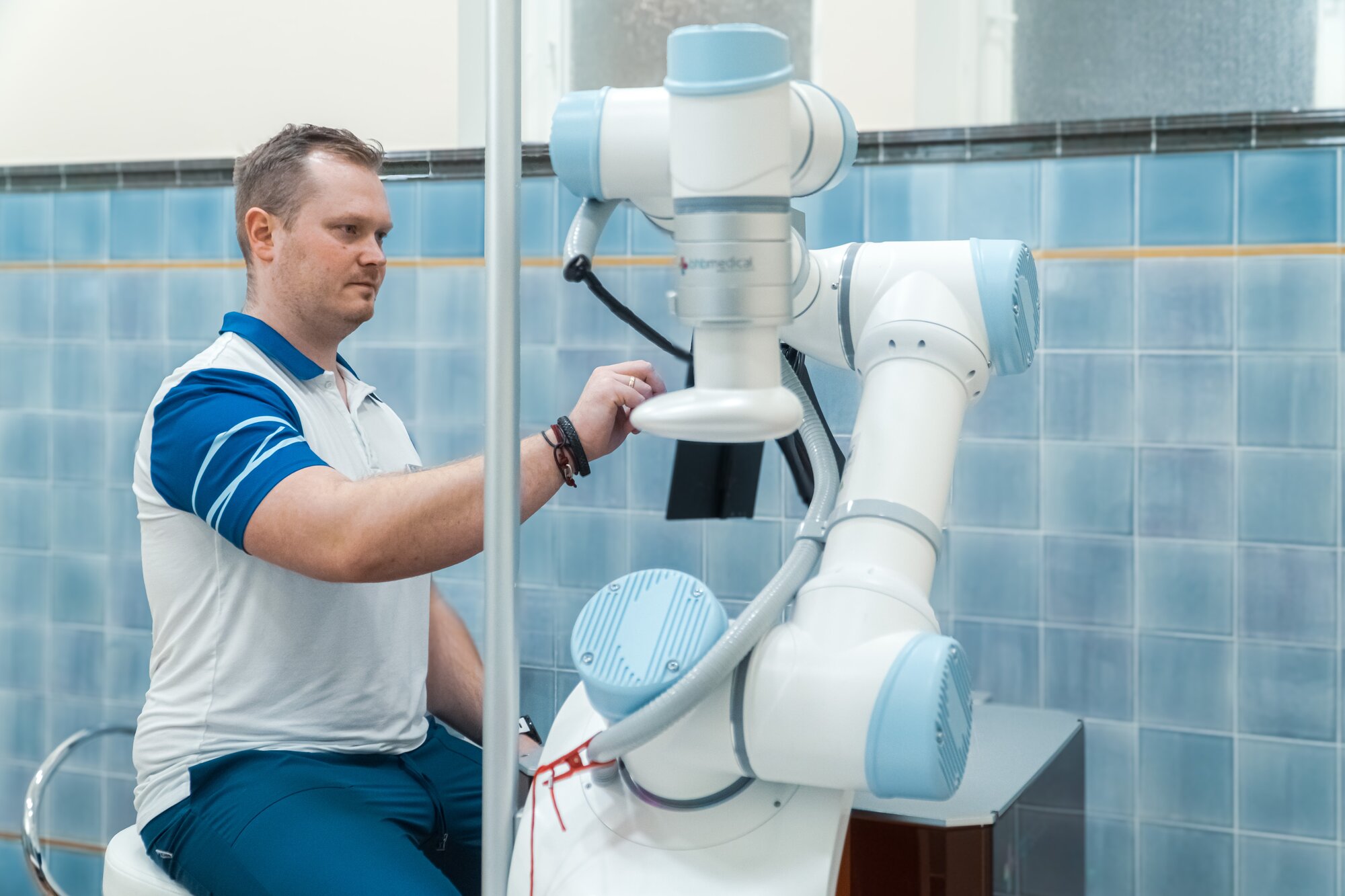Physiotherapeut am Roboterarm des Massagegeräts Rocozyer in einer Behandlungskabine mit blauen Wandfliesen