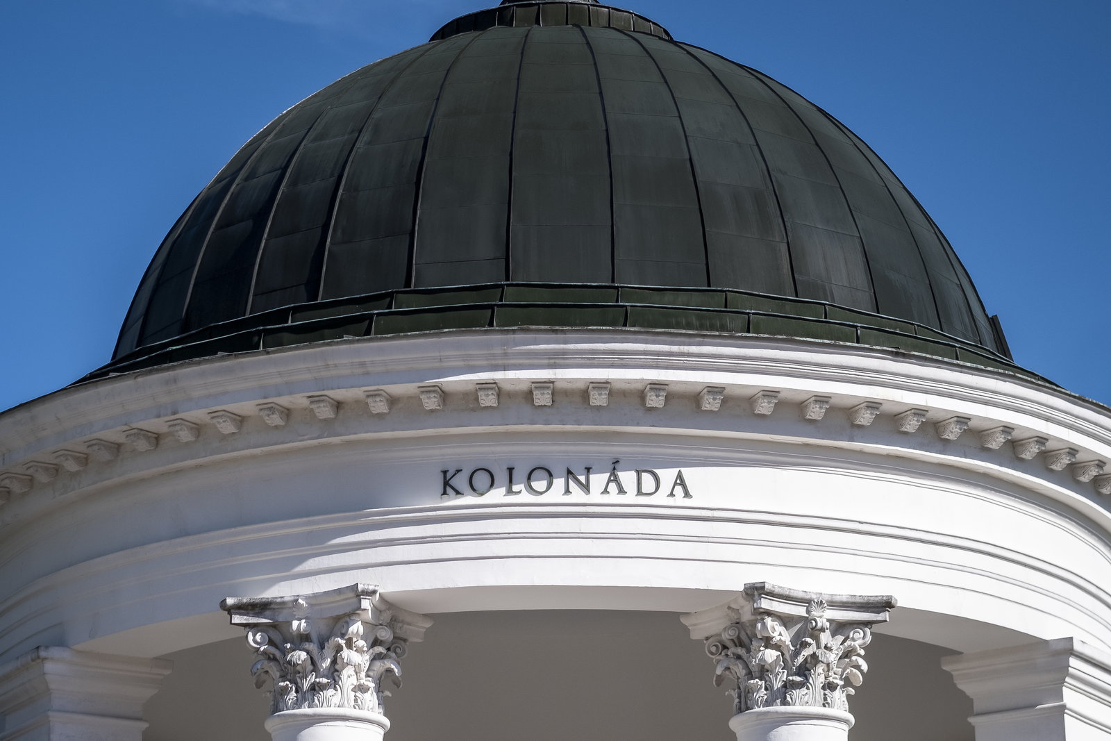Фрагмент тёмного купола и надписи Kolonáda над белым карнизом и коринфскими колоннами