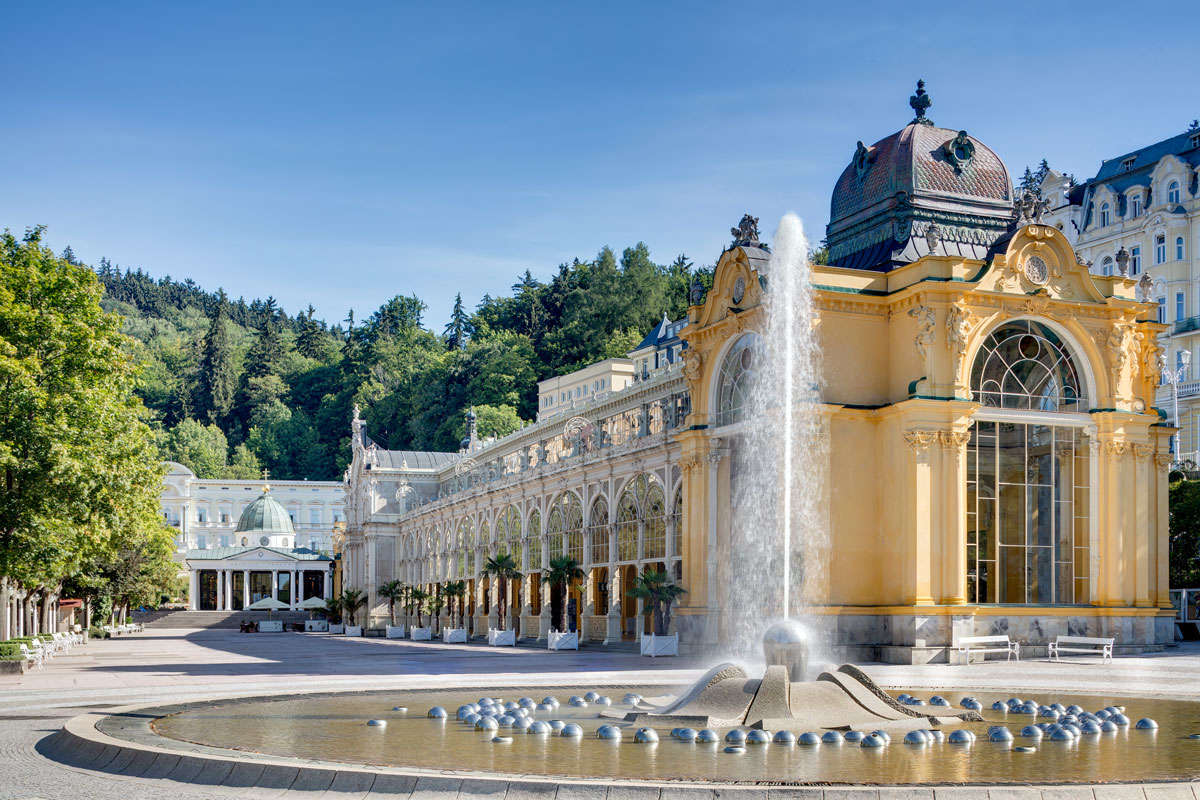 Die Singende Fontäne vor der gelben Hauptkolonnade von Marienbad, dahinter bewaldete Hügel