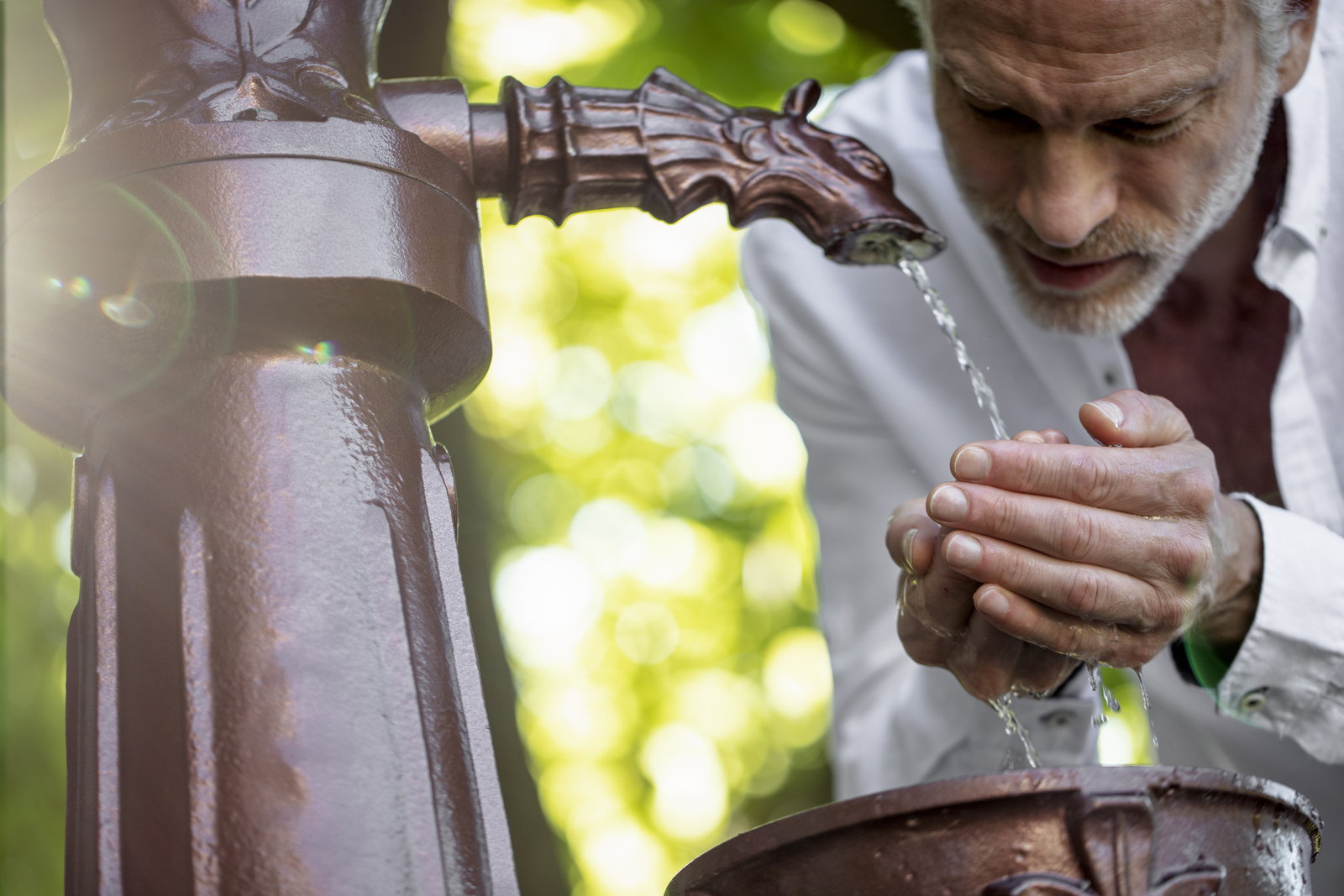 Older man cupping his hands to drink mineral water from an ornate bronze spring spout outdoors.