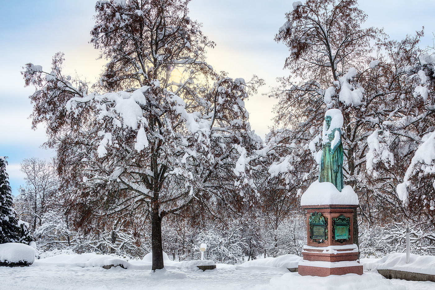 Schneebedeckte Bäume im Kurpark, rechts ein Denkmal mit grünlicher Bronzefigur auf rotem Sockel