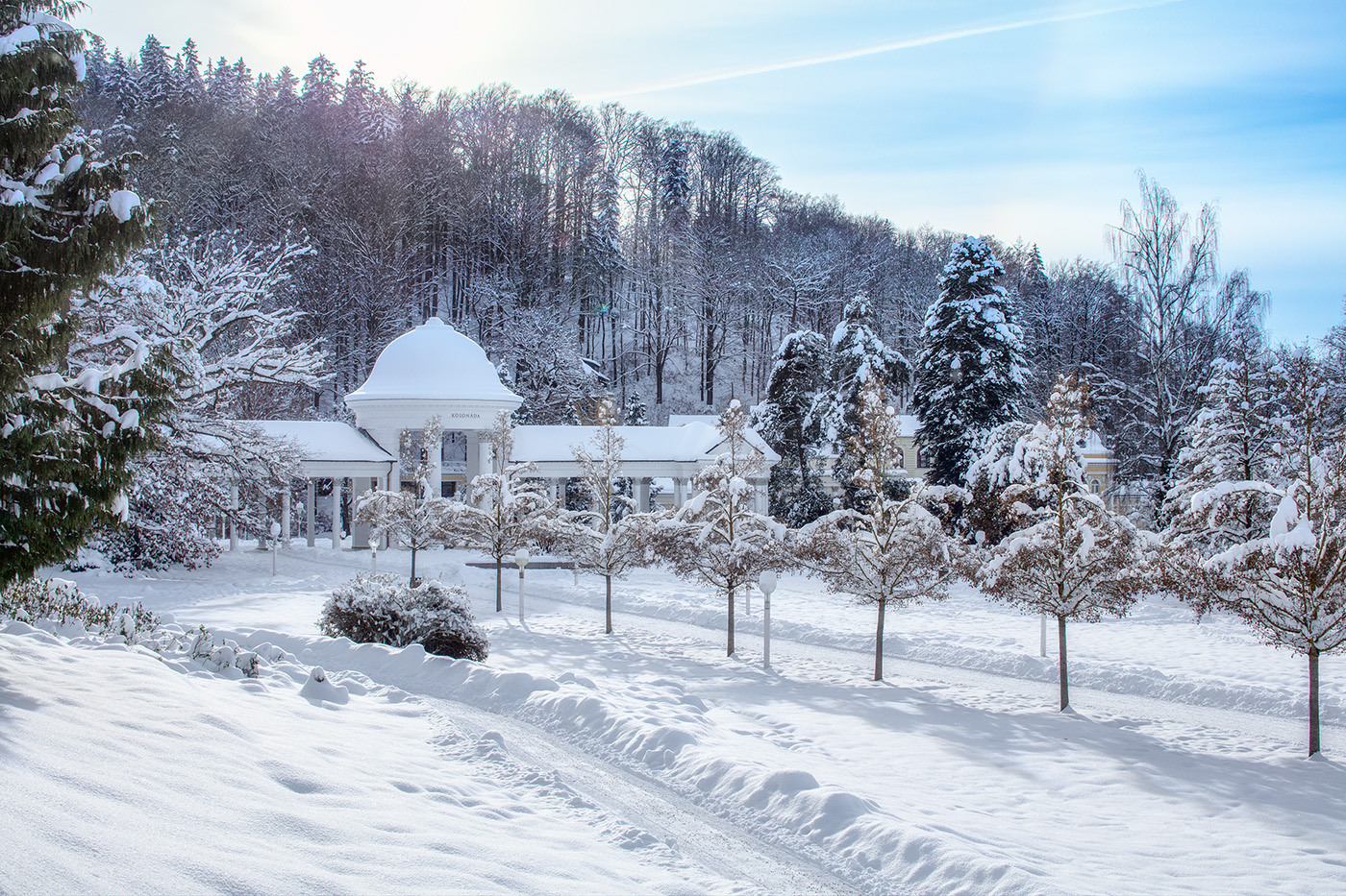 Verschneite Säulenhalle im Kurpark vor einem bewaldeten Hang, im Vordergrund Spuren im Schnee