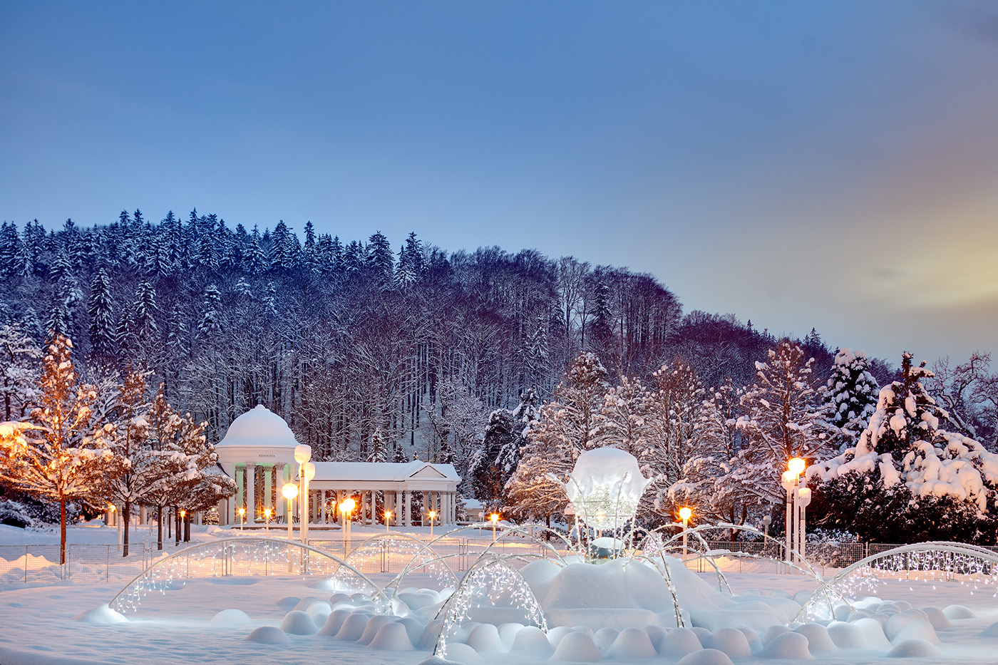 Winterabend im Kurpark: beleuchtete Bäume und Wasserstrahlen der Singenden Fontäne vor einer Säulenhalle im Schnee